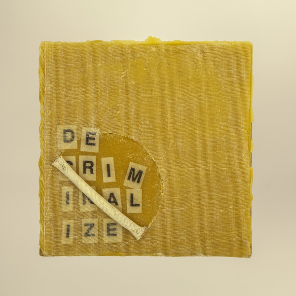 Artwork by Robin Hill that is a wooden square with beeswax, rolled cheescloth rope and letters that spell the word DECRIMINALIZE