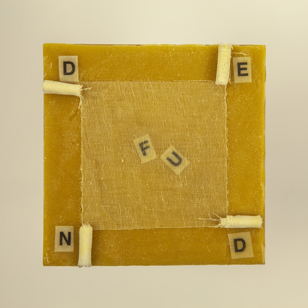 Artwork by Robin Hill that is a wooden square with beeswax, rolled cheescloth rope and letters that spell the word DEFUND