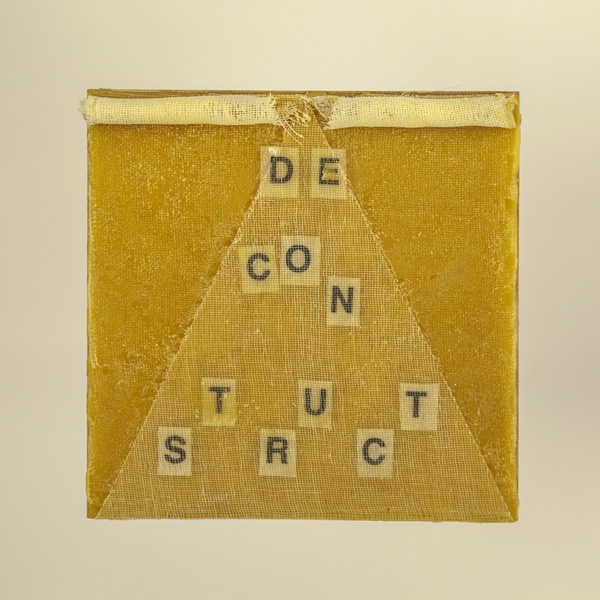 Artwork by Robin Hill that is a wooden square with beeswax, rolled cheescloth rope and letters that spell the word DECONSTRUCT