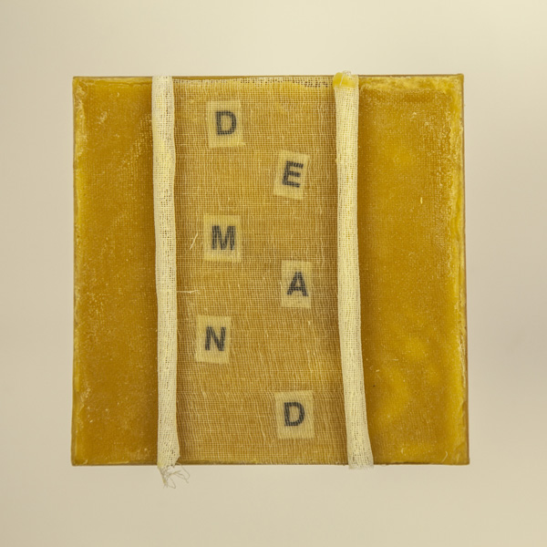 Artwork by Robin Hill that is a wooden square with beeswax, rolled cheescloth rope and letters that spell the word DEMAND