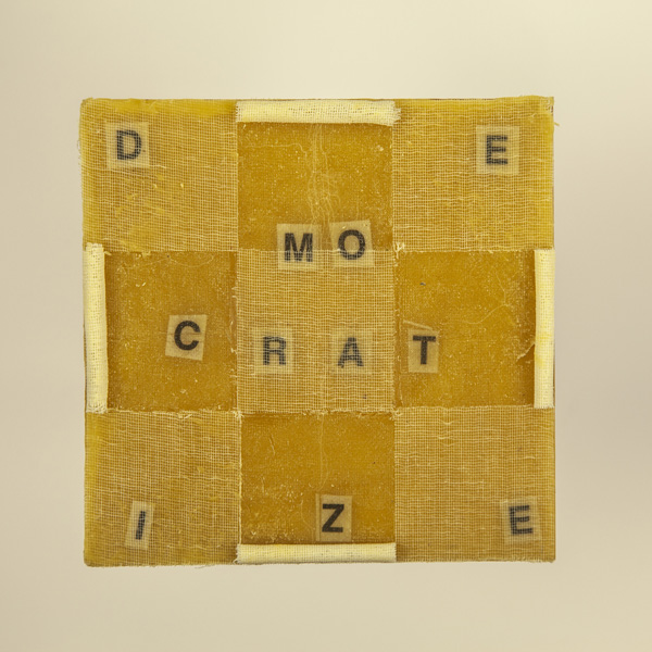 Artwork by Robin Hill that is a wooden square with beeswax, rolled cheescloth rope and letters that spell the word DEMOCRATIZE
