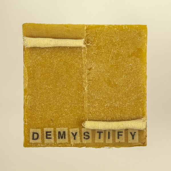 Artwork by Robin Hill that is a wooden square with beeswax, rolled cheescloth rope and letters that spell the word DEMYSTIFY