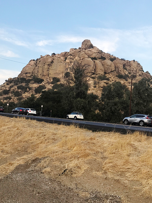 This is an image of a photograph by Vincent Johnson showing a large rock foundation with cars going by on a road in front of it and dry grass on the other side of the road